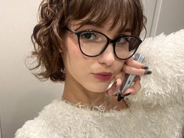 Foto em close-up de ErynAttridge, com cabelo castanho encaracolado, óculos de aro preto e um casaco branco e felpudo. Ela está segurando um telefone no ouvido, com uma expressão levemente sedutora. As unhas dela são pretas..