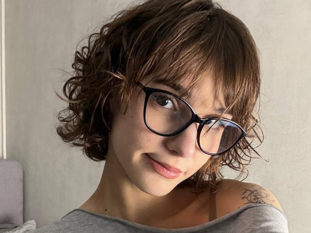 Primo piano di ErynAttridge con capelli castano ricci, che indossa occhiali con montatura nera e una maglia grigia a spalle scoperte, mostrando una un leggero sorriso, un tatuaggio discreto sulla spalla destra, una luce naturale soffusa. Che carino!.