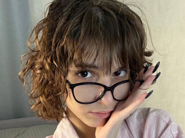 Close-up photo of ErynAttridge with messy brown hair, black-framed glasses, and long black nails, touching face with right hand, wearing a white shirt. Sunlight highlights face, creating shadow on glasses.