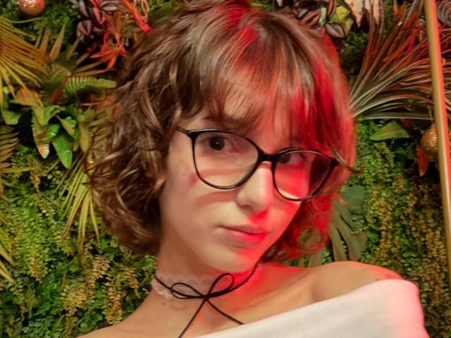 Close-up foto van ErynAttridge met rommelig bruin haar, rode highlights en een bril met zwarte montuur. Rood licht verlicht de linkerzijde van het gezicht, ze draagt een witte top met open schouders en een zwarte choker. Achtergrond: weelderige groene planten.