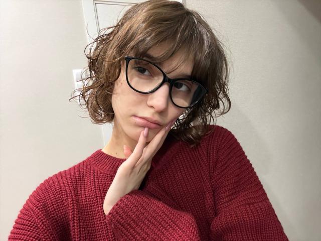 Eryn Attridge, con el pelo castaño despeinado y gafas de montura negra, lleva un suéter rojo de punto. Ella se toca la barbilla con su dedo índice, irradiando tensión sexual. Fondo blanco, iluminación brillante..