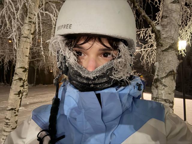 Close-up foto van ErynAttridge met een witte skihelm en een blauwe winterjas, waarvan het gezicht gedeeltelijk bedekt is met vorst. Staand in een met sneeuw bedekt bos, met kale berkenbomen. Vreselijk koud..