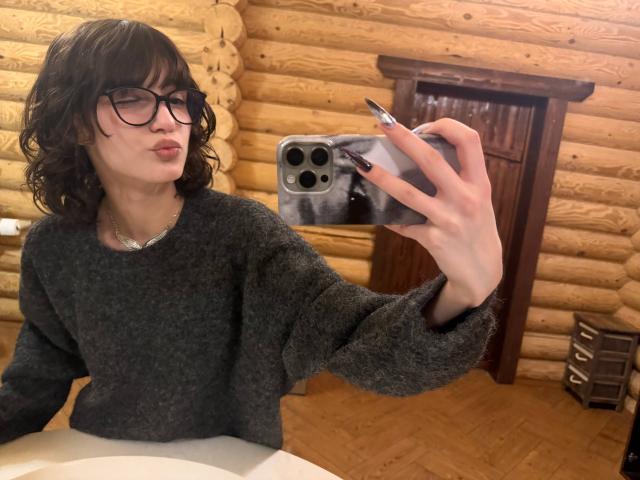 ErynAttridge, een vrouw met golvend zwart haar en een bril, maakt een selfie in een kamer van een blokhut. Ze draagt een zwarte trui en houdt een smartphone vast met een zwarte hoes. Heel aantrekkelijk..