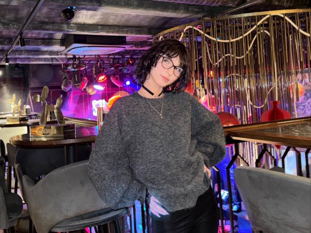 Eryn Attridge, una donna con i capelli ricci e gli occhiali, è appoggiata a uno sgabello del bar in un locale poco illuminato, con luci al neon. Indossa un maglione grigio, con una mano in tasca, emanando una forte tensione sessuale..