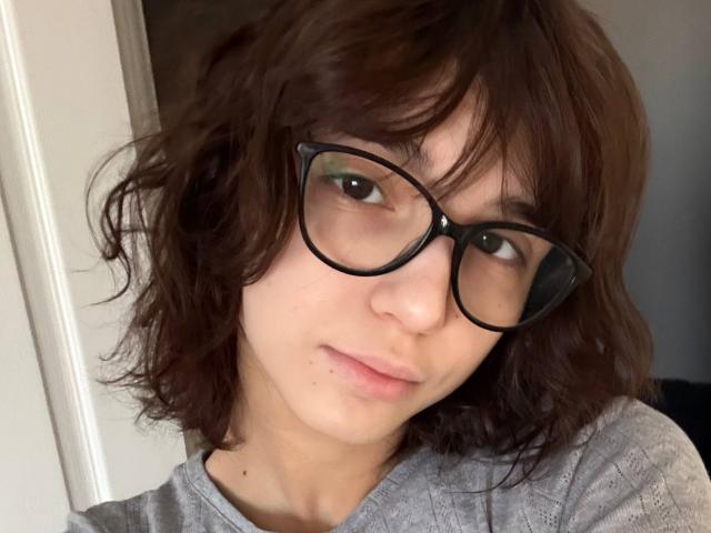 Primer plano de ErynAttridge con cabello castaño despeinado, gafas de montura negra y una expresión neutra. Ella lleva una camisa gris, y su piel clara contrasta con el fondo oscuro. Increíblemente atractivo/a..