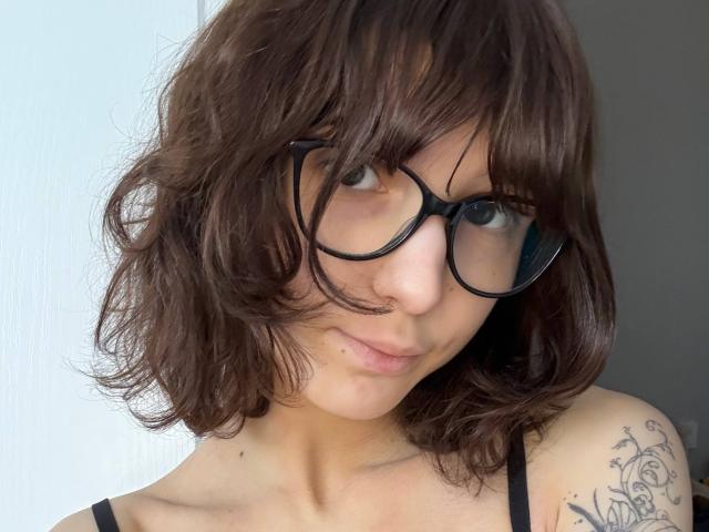 Portrait rapproché d'ErynAttridge, avec des cheveux bruns ondulés, des lunettes à monture noire et un tatouage sur l'épaule droite. Elle regarde directement l'appareil photo, et elle porte un haut noir à bretelles fines. Tellement mignon..