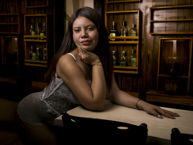 NatashaSx, met lang donker haar, leunt verleidelijk over een houten bar, gekleed in een strak grijs kanten topje, met haar hand onder haar kin. donkerhuidig, flessen op houten planken op de achtergrond, verleidelijke uitdrukking.