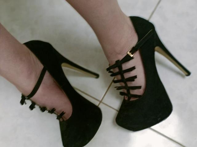Close-up of WollyMolly's legs in black high-heeled shoes with lace-up straps, standing on white tiled floor. Shoes have gold buckles, heels are gold-tipped. Legs are pale, feet slightly angled.