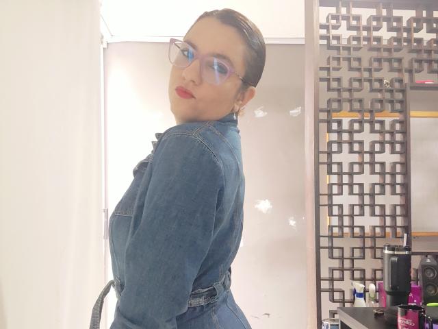 Amelia Wilson, met haar haar naar achteren gekamd, draagt een denim shirt, blauwe oogschaduw en rode lippenstift, en kijkt over haar schouder. schouder. Lichte achtergrond, geometrische muur, schoonheidsproducten aan de rechterkant. Heel aantrekkelijk..