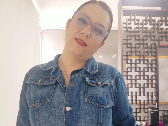 Amelia Wilson, con i capelli castano scuri pettinati all'indietro, indossa una camicia di jeans blu e occhiali trasparenti, ha un leggero sorriso e i capelli rossi. rossetto. Lo sfondo mostra una moderna cantina. Incredibilmente sexy..
