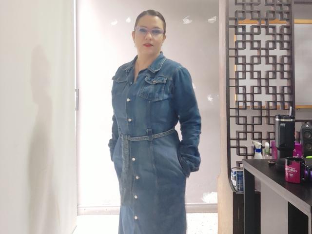 AmeliaWilson stands confidently in a bright room, wearing a blue denim button-up dress with pockets, hands in her pockets. She has red lipstick and glasses, next to a shelf with pink bottles.