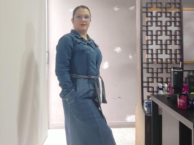 Amelia Wilson está de pé, com confiança, em uma sala bem iluminada, vestindo um vestido de jeans azul, com as mãos nos bolsos e óculos. e uma expressão séria. Do lado direito, há prateleiras pretas com garrafas cor-de-rosa..