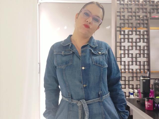 Amelia Wilson è in piedi in una stanza ben illuminata, indossa un vestito di jeans blu con una cintura e occhiali trasparenti. Il suo rossetto rosso contrasta con la sua pelle chiara. Lo sfondo presenta una parete geometrica e prodotti di bellezza. Molto attraente..