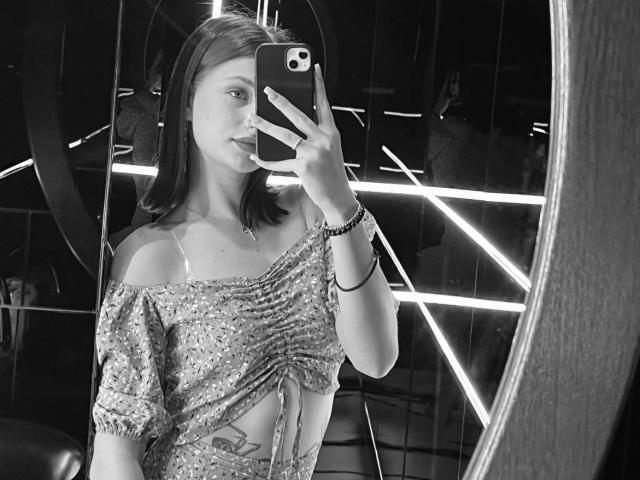 Black-and-white photo of CleliaMarchak taking a mirror selfie. She's wearing a sparkly off-shoulder top, holding her phone with her left hand, exposing her cleavage. Neon lights in background.