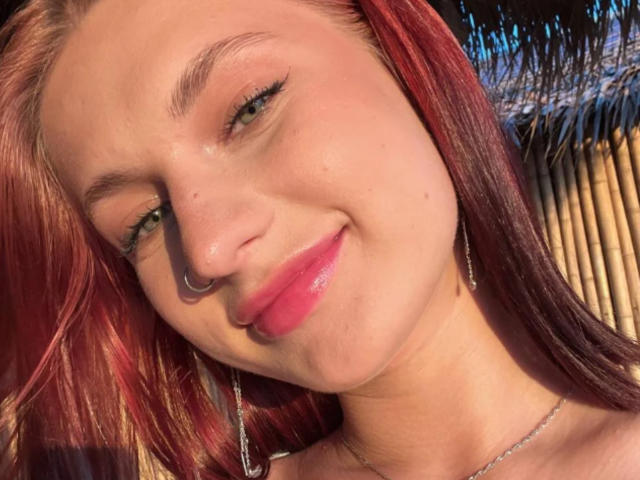 Close-up of CleliaMarchak with red hair, wearing a nose ring and dangling earrings, smiling with pink lipstick. Sunlight highlights her face, creating sharp shadows and a sensual glow.