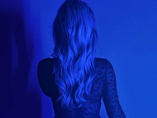 BalletSwan, facing away, bathed in blue light, long wavy blonde hair, sheer black top, visible back and shoulders, silhouette, strong sexual tension, hair cascading down, intense blue glow.