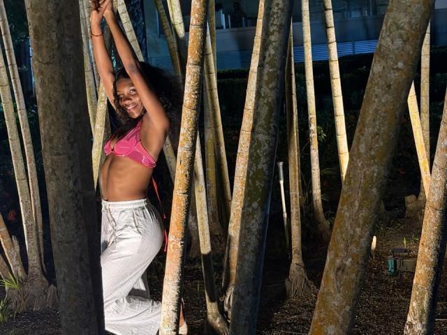Uma fotografia de Alanha em pé, cercada por árvores altas e finas. Alanha tem a pele escura, cabelos longos, e está vestindo um top esportivo rosa e calças brancas, com os braços levantados, sorrindo, e com a barriga visível. abs.