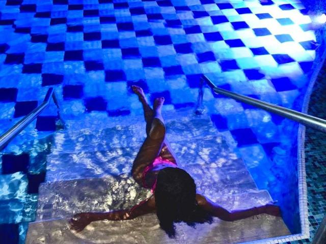 Ein Foto von Alanha mit dunkler Haut, nass und teilweise im Wasser eines blau gefliesten Pools, mit gespreizten Beinen und einem hochgehobenen Arm. eine sinnliche, erotische Atmosphäre schaffen, mit sichtbaren Wasserspritzern..