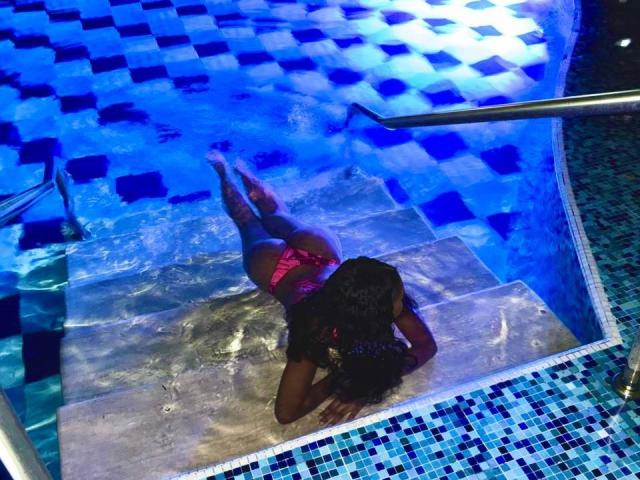 Una fotografía de Alanha acostada boca abajo en una piscina iluminada con luz azul, vistiendo un bikini rojo, con una pierna levantada, y el agua ondulando. alrededor de ella. Azulejos azules brillantes e iluminados rodean la piscina..