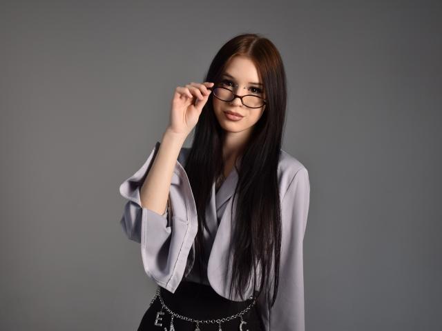 Abigail Morris, uma mulher magra com cabelos longos e pretos, vestindo uma blusa cinza e um vestido preto, ajusta seus óculos pretos. Mão direita. Tensão sexual, sorriso discreto, olhos escuros..