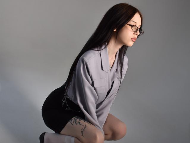 Abigail Morris, met lang zwart haar en een bril, knielt in een grijze blouse, een zwarte rok en witte sokken. Ze heeft een tatoeage op haar dij, die verleidelijk naar de zijkant kijkt..