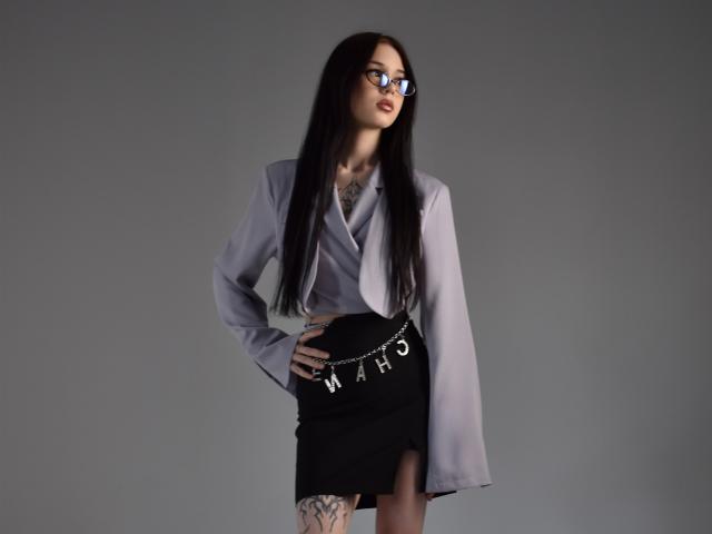 AbigailMorris with long black hair, wearing glasses, a loose gray shirt, and a black skirt with a belt. She stands with one hand on her hip, looking to the side. Fucking sexy.