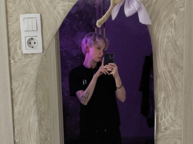 Dionne Szczygiel, uma pessoa magra, com tatuagens e cabelo loiro curto e desgrenhado, está em frente a um espelho, tirando uma selfie. com um telefone preto. Usando uma camisa preta, com iluminação roxa. Incrivelmente sexy..