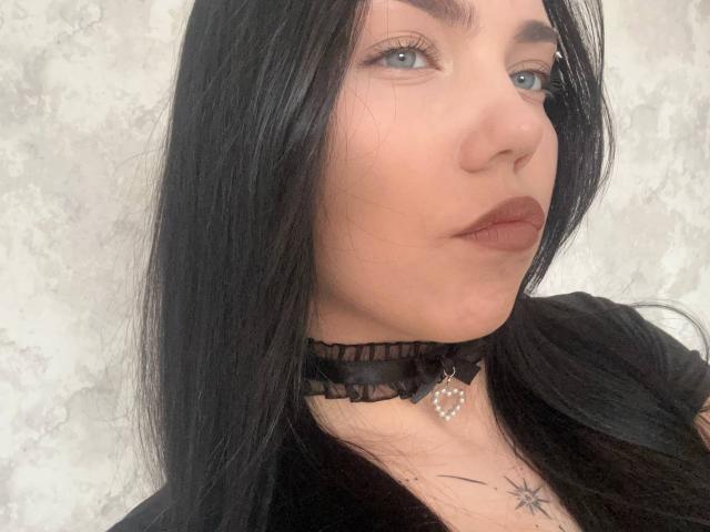 Close-up de Ghislaine Koplin, com pele clara, cabelo preto e olhos azuis. Ela usa um colar preto de renda com um pingente de coração, e tem uma pequena tatuagem de estrela no pescoço. Muito atraente..