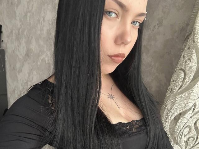 Photo en gros plan de Ghislaine Koplin, avec de longs cheveux noirs, une peau pâle et des yeux bleus. Elle porte un haut en dentelle noire, qui révèle un tatouage sur sa poitrine. La tension sexuelle est palpable..