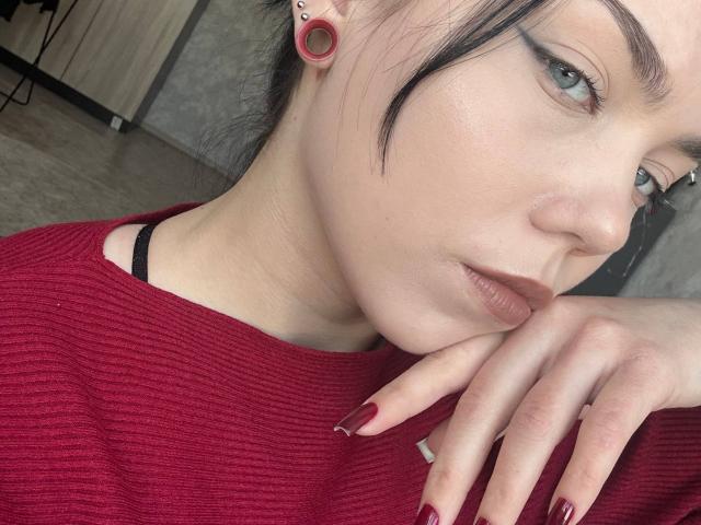 Photo en gros plan de Ghislaine Koplin, une femme blanche à la peau pâle, aux cheveux noirs, avec des piercings rouges aux oreilles et un vernis à ongles rouge. Elle regarde de côté, sa main près de son visage, et porte un pull rouge..