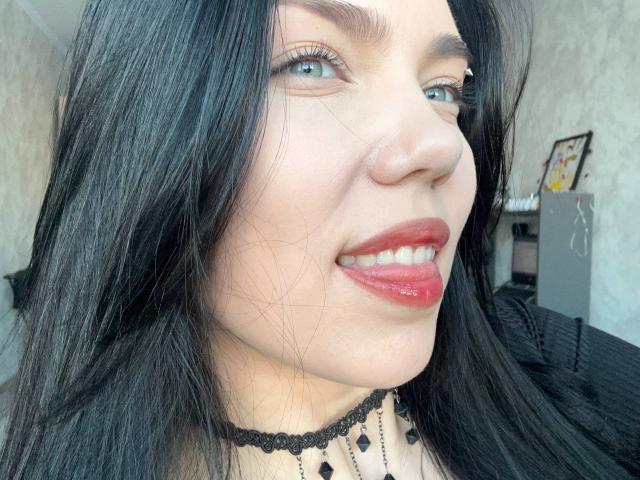 Close-up van Ghislaine Koplin met lang zwart haar, helderblauwe ogen en rode lippenstift. Ze kijkt naar de zijkant met een lichte glimlach. Haar choker heeft zwarte diamantpatronen. De achtergrond toont een wazige kamer..