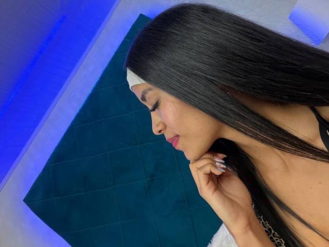 Close-up photo of LaurentRay with long black hair, wearing a white headband, gazing downward with a slightly sultry expression. Blue and black background, hand touching chin. Sexual tension, sexy vibe.