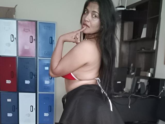 ArianithaXox, a curvy woman with long black hair, gazes over her shoulder, biting her finger. She wears a red bra and black skirt, with colorful lockers in the background. Fingers near mouth.