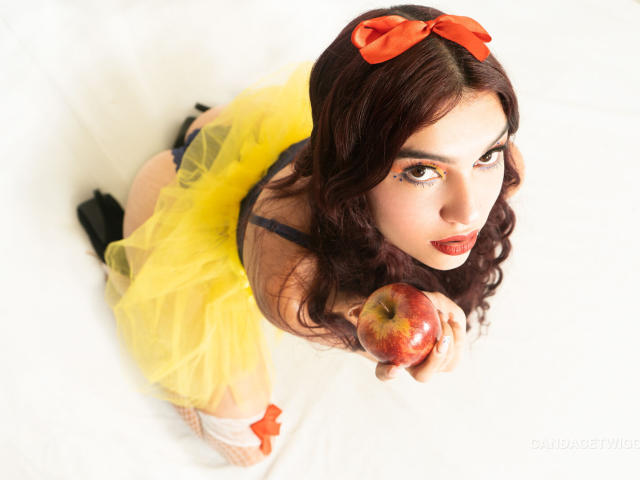 Gros plan de Noemi Ferrer, avec des cheveux noirs ondulés, un nœud rouge, une robe en tulle jaune, tenant une pomme rouge. Elle lève les yeux, les lèvres légèrement entrouvertes, d'une manière sexy et séduisante. Fond blanc éclatant.