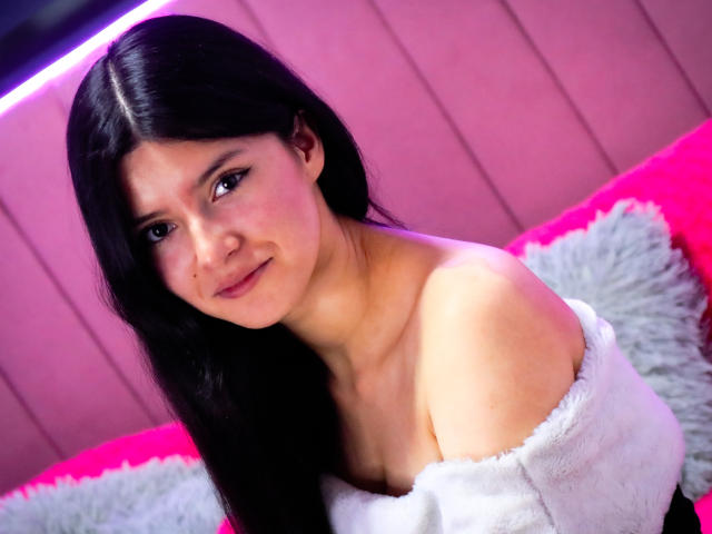 EmiSchwarz, with long black hair and light skin, gazes directly at the camera. She's wearing a white off-shoulder top, exposing her collarbones. Background includes pink and white textures. Fucking sexy.