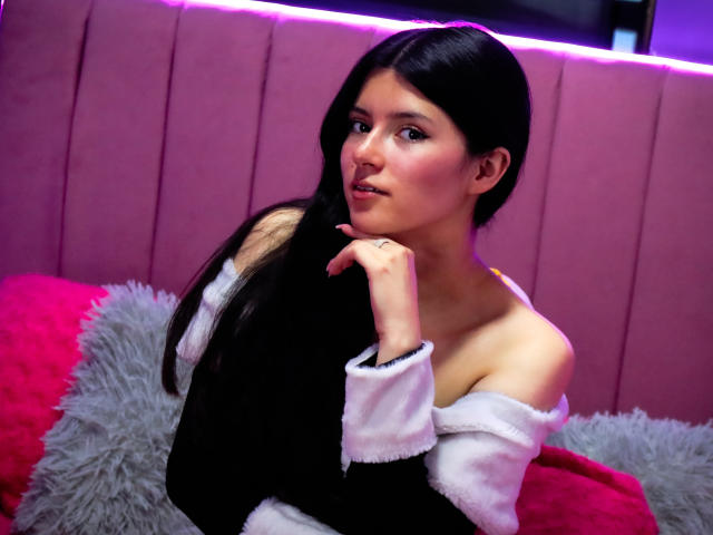 EmiSchwarz with long black hair, sexy expression, and white off-shoulder top, touching her chin. Pink and red pillows, gray fluffy pillow in the background. Neon purple strip lights above.