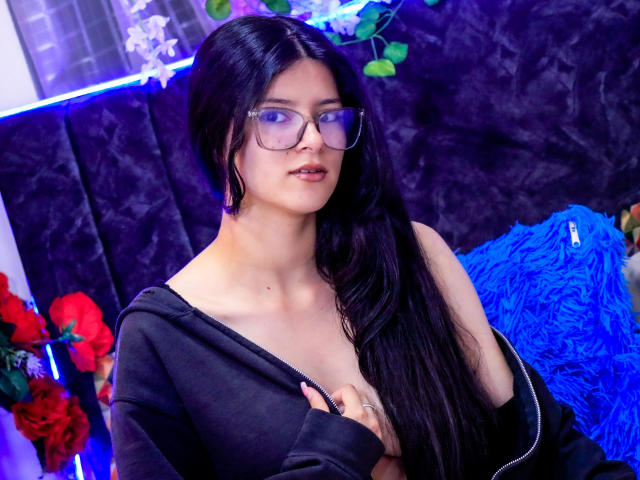 EmiSchwarz, con cabello negro largo, usando gafas y una blusa negra con un escote pronunciado, que deja ver parte de su pecho. Ella está mirando a la cámara con la boca ligeramente abierta, y hay una iluminación azul en el fondo..