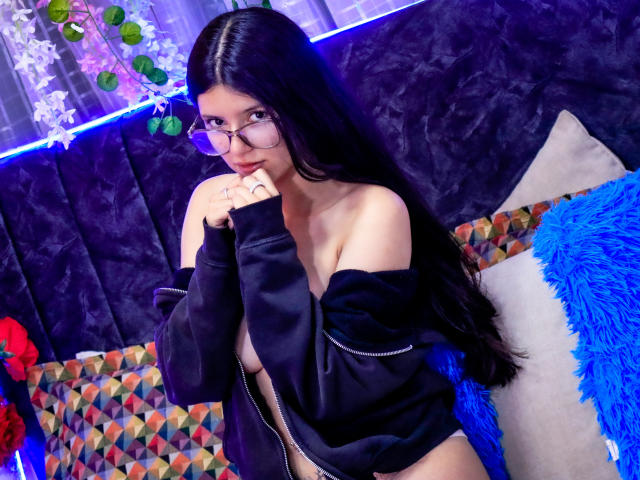 EmiSchwarz, a slim woman with long black hair and glasses, wears an off-shoulder black top, biting her finger seductively. She's on a couch with colorful pillows, blue and white fur. Fucking sexy.