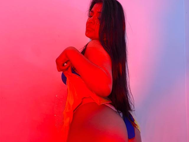 ArianithaXox, avec ses cheveux noirs mouillés, se tient de profil, vêtue d'un maillot de bain bleu. Une lumière néon rouge et rose illumine son corps aux courbes généreuses et voluptueuses. Sa main repose sur sa hanche, dégageant une tension sexuelle..