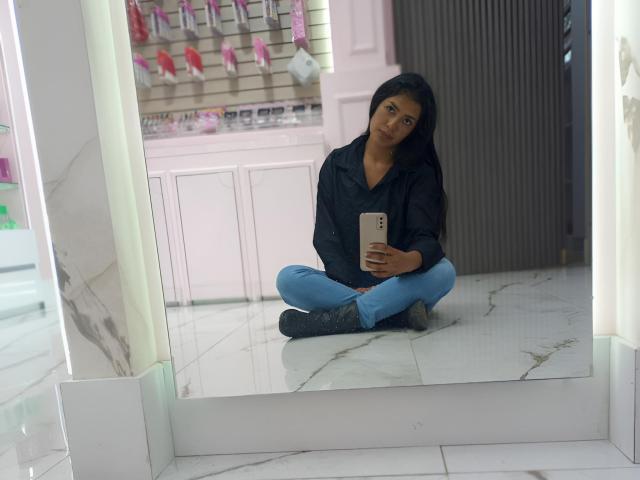 ArianithaXox, une femme aux formes généreuses avec de longs cheveux noirs, est assise les jambes croisées sur un sol en marbre blanc, vêtue d'une veste noire et jean bleu, tenant un smartphone, le regardant de manière séduisante. Bien éclairé.