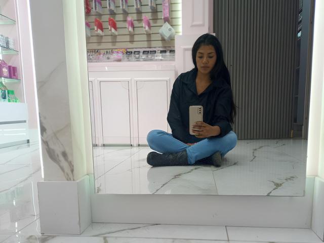 ArianithaXox, with dark hair, sits cross-legged on a white marble floor, wearing a black shirt and blue jeans, holding a phone, reflecting in a mirror, surrounded by brightly lit shelves.