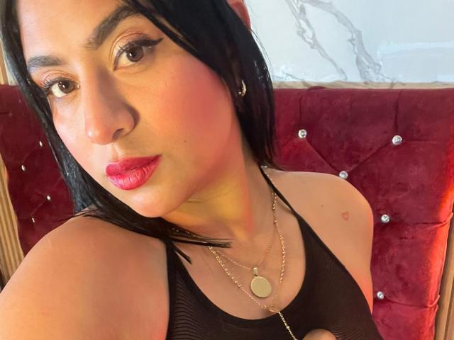 Close-up van DiannaRosse, een Latina vrouw met zwart haar, rode lippenstift en donkere eyeliner, die een zwarte top van gaas draagt. Gouden ketting. Ze zit op een rode, gestoffeerde bank en kijkt verleidelijk in de camera..