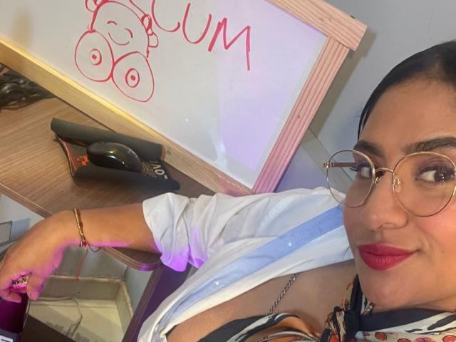 DiannaRosse, con cabello oscuro y lápiz labial rojo, se inclina hacia adelante, vestida con una camisa blanca que deja entrever un poco de escote, y lleva gafas. Pulsera de oro.
el fondo incluye un letrero de neón con letras rojas cursivas..