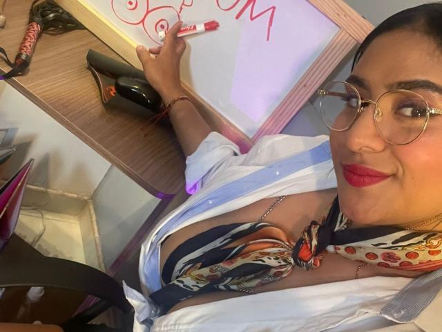 DiannaRosse met rode lippenstift, een bril en een wit shirt dat open is om de decolleté te laten zien, schrijft met rode stift op een whiteboard. markering. Ze draagt een kleurrijke sjaal en heeft een zelfverzekerde glimlach..