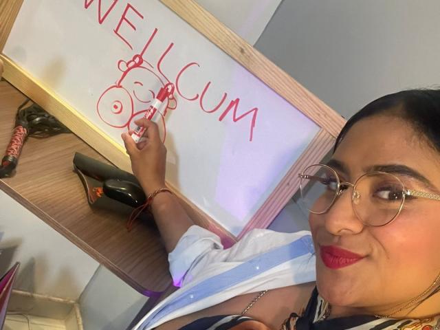 DiannaRosse mit Brille und rotem Lippenstift, die mit einem roten Marker "WILLKOMMEN IN CUM" auf einer Whiteboard schreibt. Sie hält ein Messer, auf dem Tisch befinden sich eine Flasche Wein und ein schwarzer Stift..