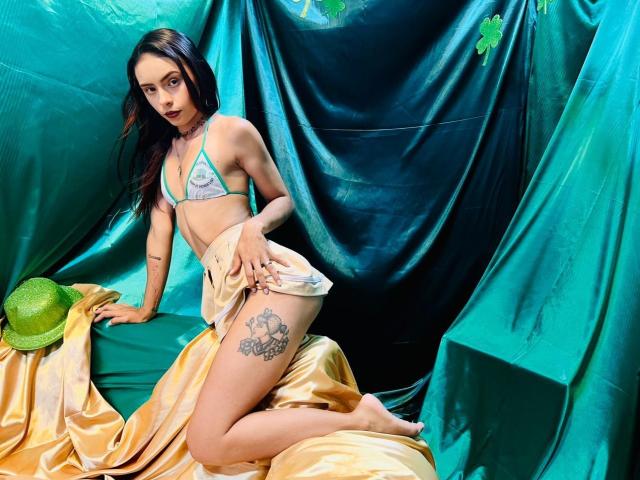 ValeryHotLoove, arrodillada sobre seda dorada, vestida con un bikini azul, mostrando un tatuaje en su muslo izquierdo. Gorro verde a la izquierda, fondo de cortina negra. Pose sexy y seductora, cabello oscuro, mirada intensa..