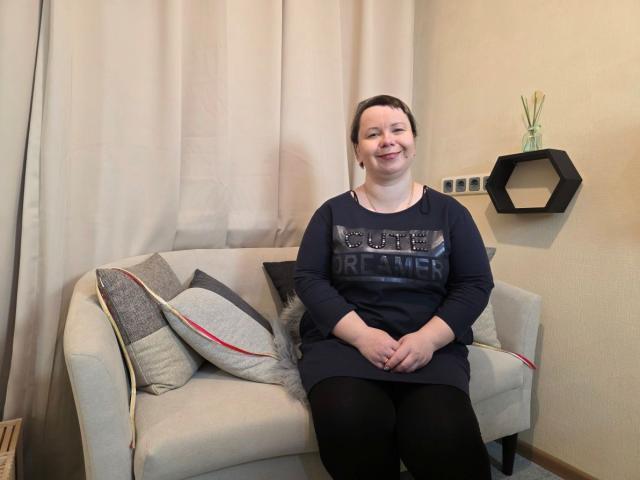 BonnnyBoobsy sits on a light beige couch, smiling, wearing a black long-sleeve shirt with a graphic design. Cushions with grey and red accents are on the couch. Hexagonal shelf with vase.