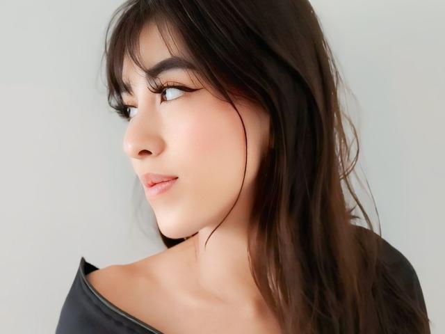 Photo en gros plan de VanellopeLm, avec des cheveux brun foncé, vue de profil, regardant vers la gauche. Elle a la peau claire, un trait d'eye-liner noir, un rouge à lèvres rose discret, et porte un haut noir. Tension sexuelle évidente..