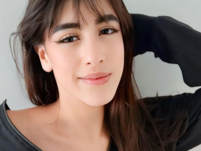 Primo piano di VanellopeLm, una giovane donna con lunghi capelli scuri, che indossa una maglietta nera e ha una mano sollevata dietro la testa. Ha la pelle chiara, gli occhi scuri e un leggero sorriso. L'illuminazione è intensa..