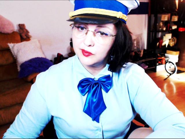 ImperatrizaSanda, con piel pálida y lápiz labial rojo, vestida con un uniforme de azafata de vuelo azul y blanco con un gran detalle azul. Un lazo, y un sombrero azul con una banda amarilla. Fondo interior borroso.